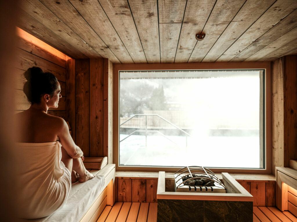 A woman sits in the sauna and gazes out at the snowy landscape. The sauna is powered by solar and hydroelectric energy.