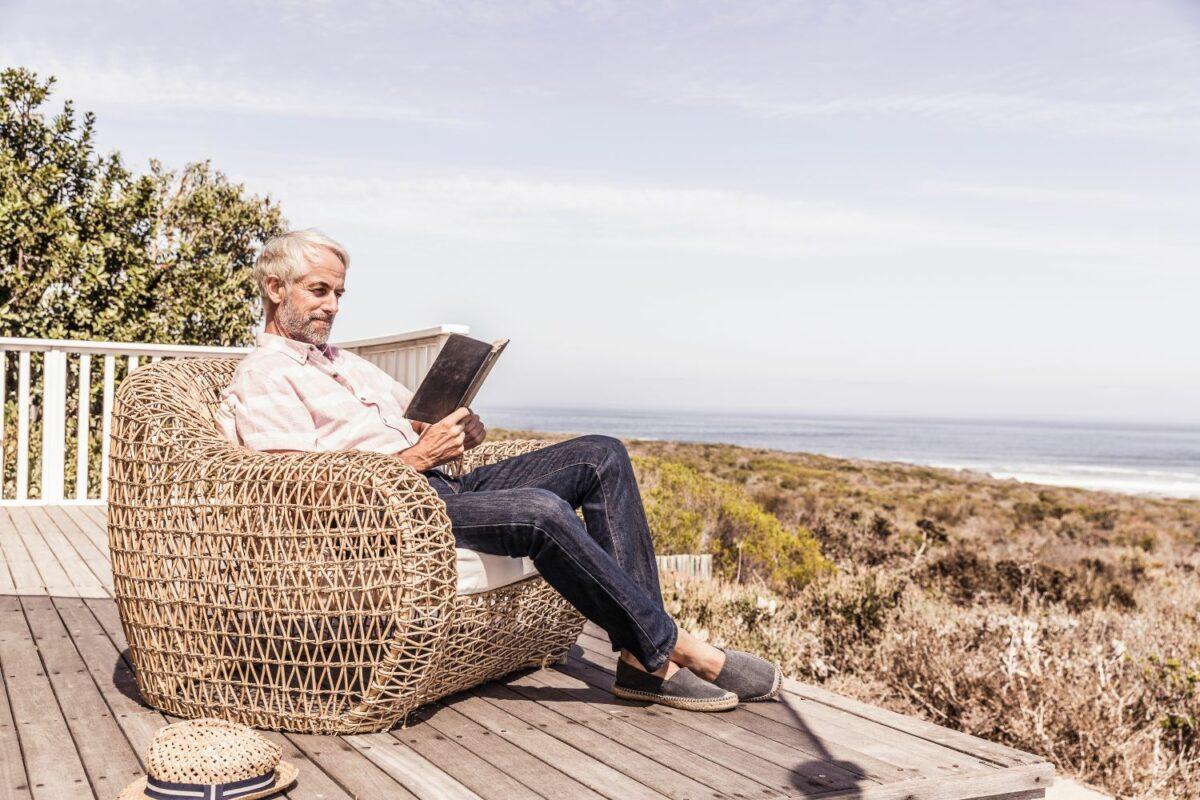 A man in a wicker chair reads a book while traveling, looking out at the beach