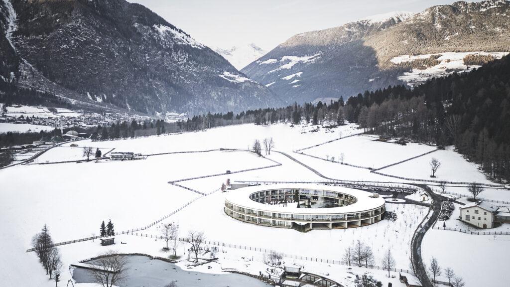 Luftaufnahme eines modernen, kreisförmigen Gebäudes, umgeben von schneebedeckten Feldern und Bergen, mit einer ruhigen Winteratmosphäre und klarem blauem Himmel.