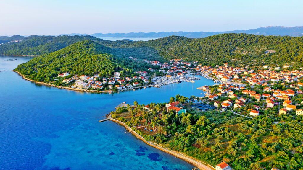 This aerial view of Iz, an island in Croatia, features colorful houses nestled between lush green hills and a clear blue bay. The scene creates a peaceful and picturesque atmosphere.