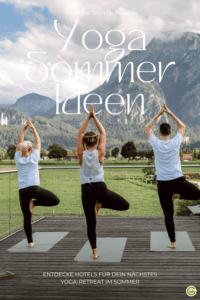 Drei Personen praktizieren Yoga im Baumstand auf Yogamatten vor einer Berglandschaft mit Wiesen und Bäumen